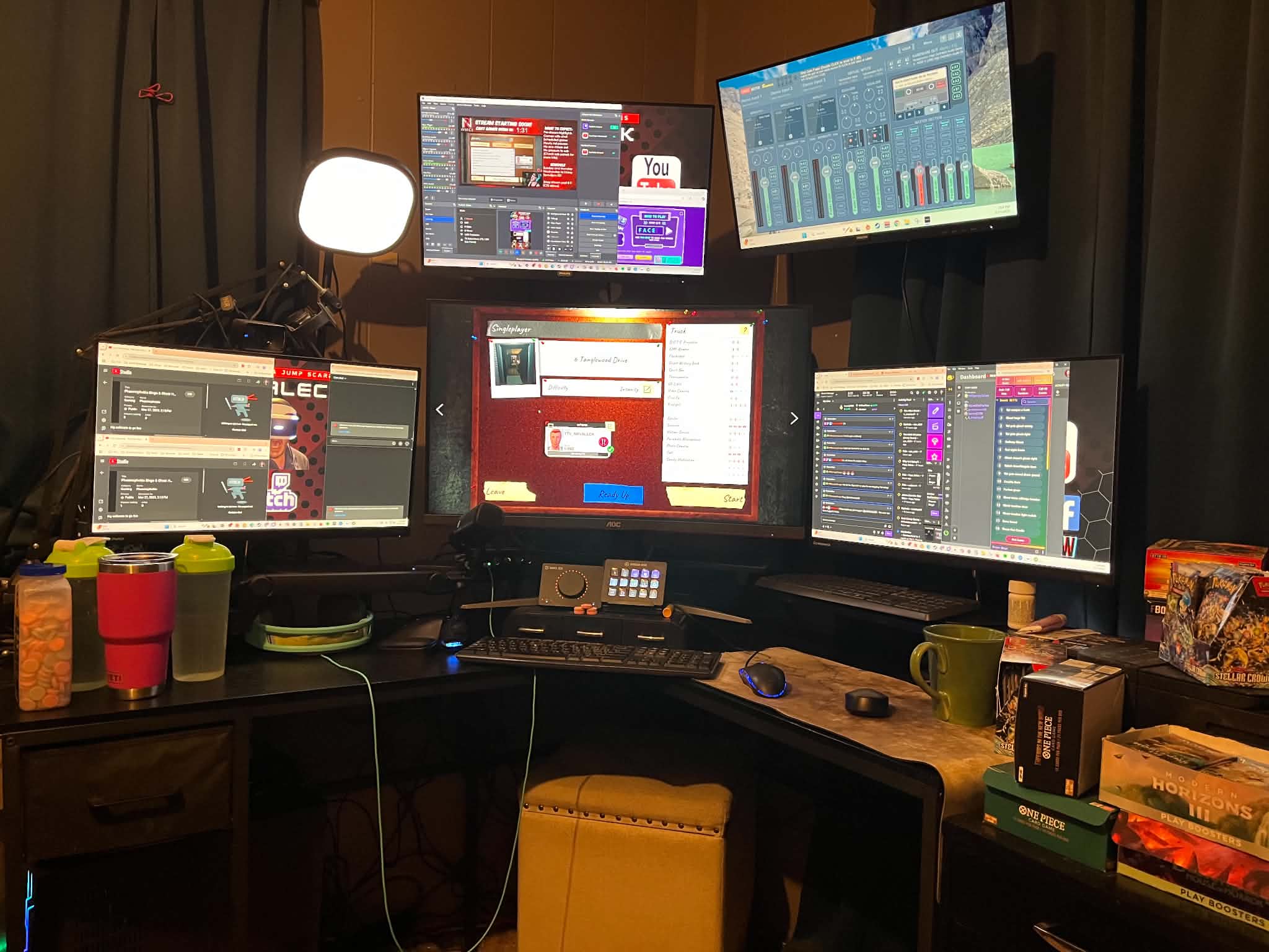 My streaming battlestation: a five-monitor layout with a central curved gaming monitor, stream dashboards, two Stream Decks, an audio mixer, and gaming peripherals on an L-shaped desk.